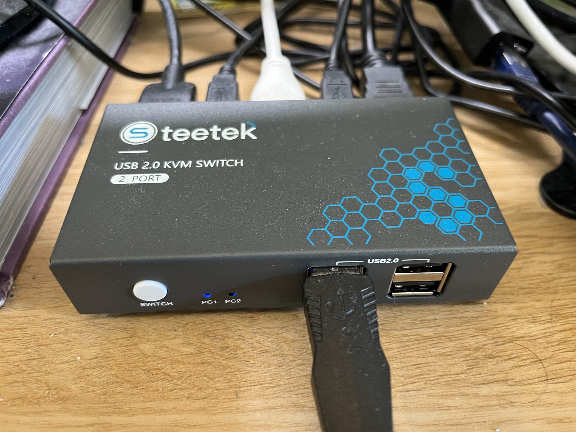 A photo of the Steetek KVM switch, with a USB cable plugged in the front and various USB and HDMI cables plugged into the back. It's a grey, rectangular metal box.